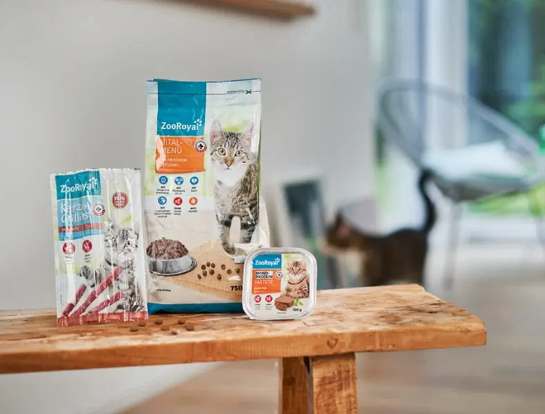 ZooRoyal Katzenfutterprodukte im Wohnzimmer ZooRoyal Katzenfutterprodukte stehen auf einer Bank, während eine Katze im Hintergrund durch das Wohnzimmer läuft.