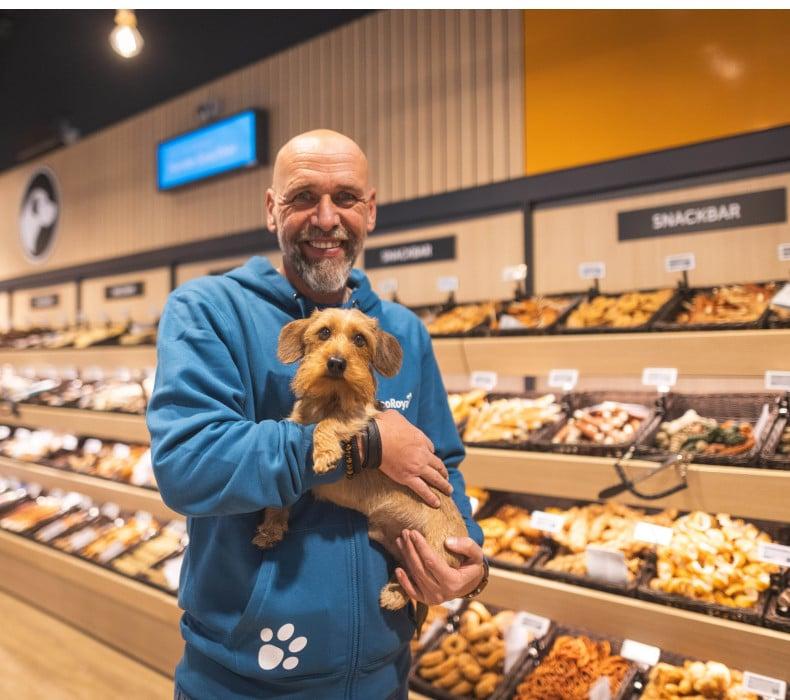 Snackregale im ZooRoyal Fachmarkt Stellingen Ein Mann hält einen kleinen braunen Hund vor Regalen mit Hundesnacks