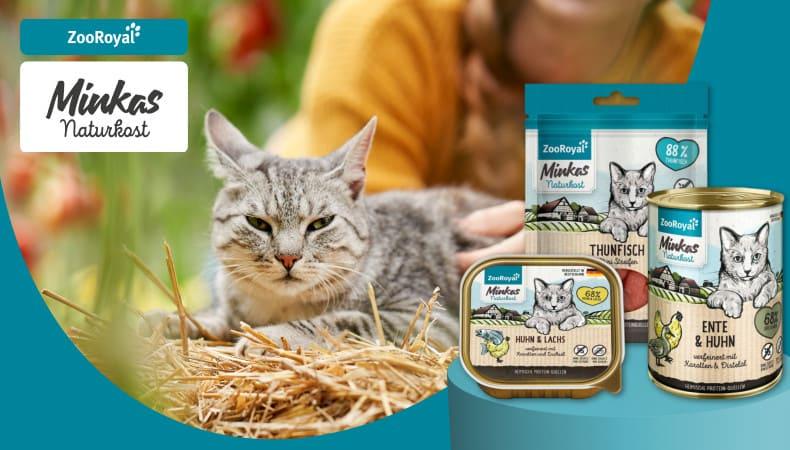 ZooRoyal Eigenmarken Minkas Naturkost Katze liegt im Stroh neben ZooRoyal Minkas Naturkost Produkten.